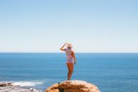 Woman in White Tank Top and Blue Denim Shorts Standing on Brown Rock Formation Near Body