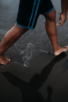 Barefoot individual stepping on wet black sand with fishing net, creating a coastal vibe.
