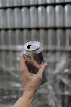 A hand holding an aluminum can in a brewery setting with rows of cans in the background.