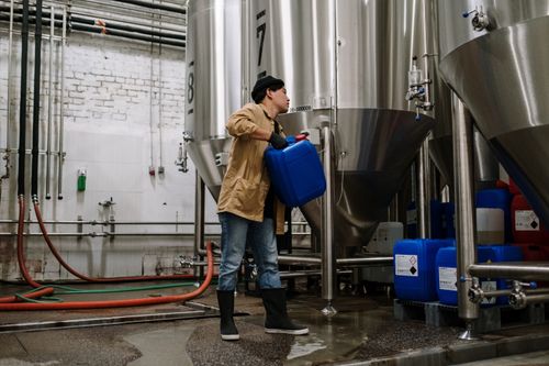Free Man handling blue container in a brewery with stainless steel tanks. Stock Photo