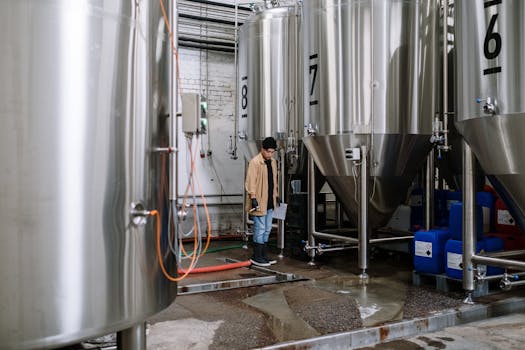 An industrial worker inspects fermentation tanks in a brewery, surrounded by equipment and machinery.