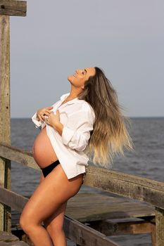 Pregnant woman stands on a wooden boardwalk by the sea, enjoying the breeze.