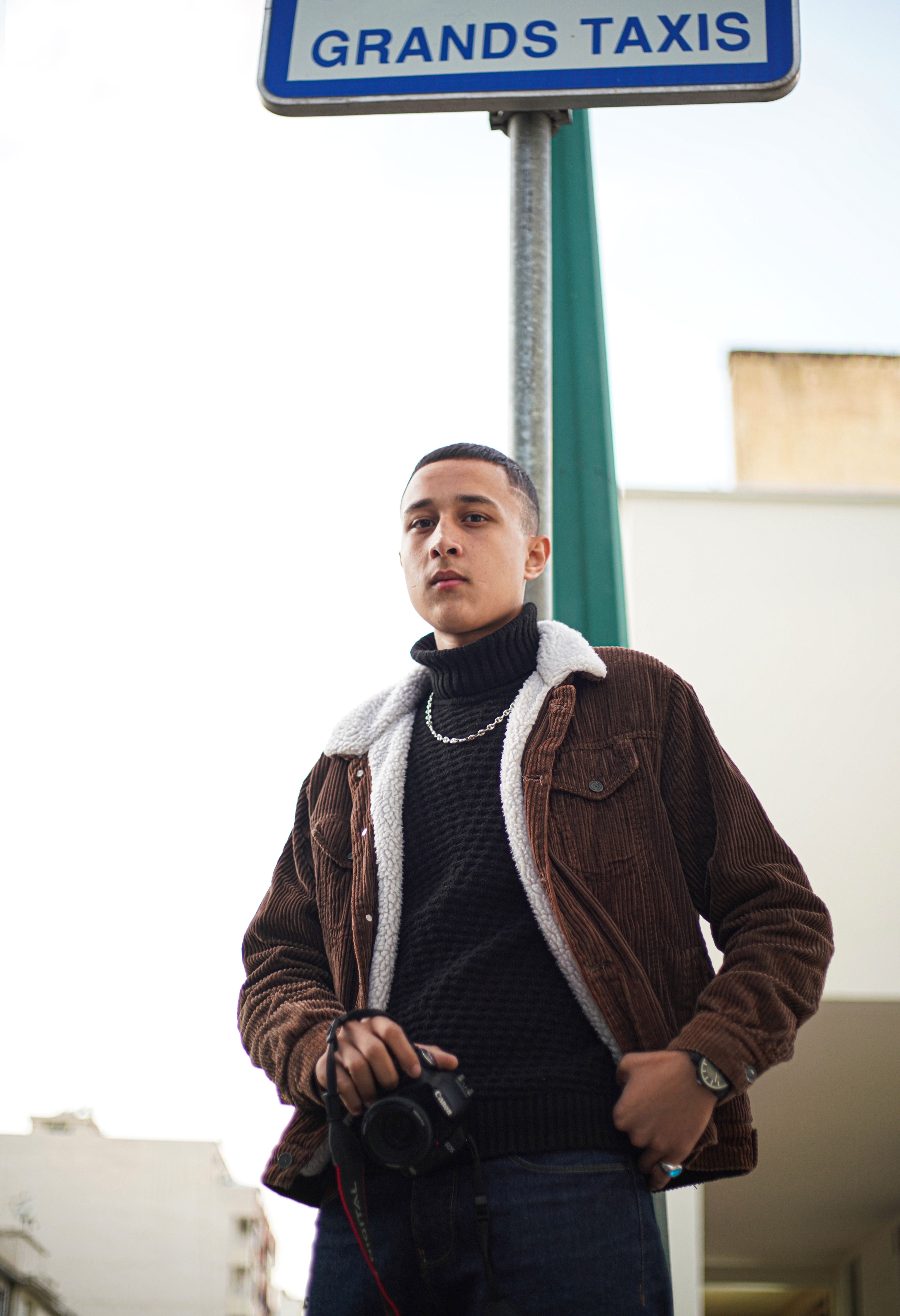 Young man in a brown jacket posing in Meknes, Morocco, holding a camera.