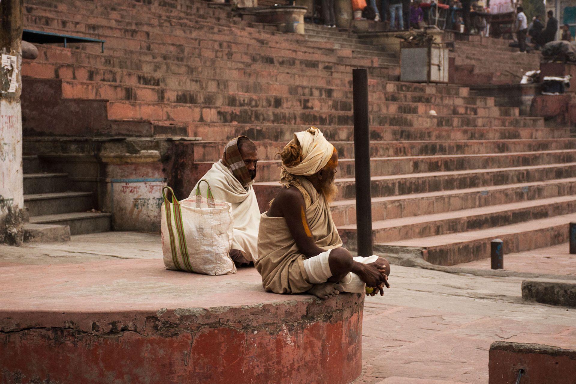 Two senior Indian men in traditional clothes sitting outdoors on stone steps in an ancient city setting.