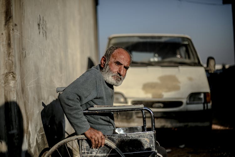 Senior Ethnic In Wheelchair Near Concrete Wall Against Car