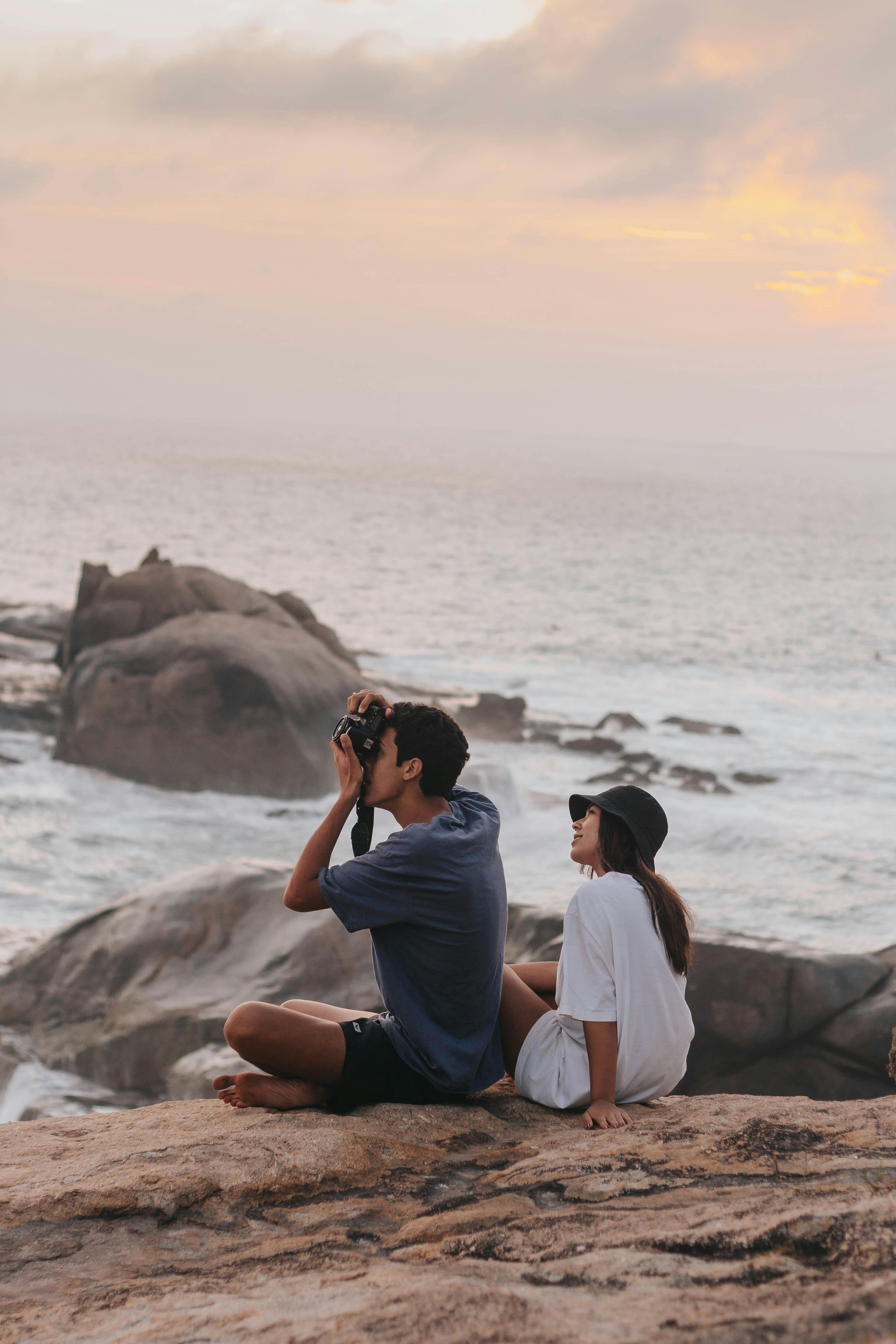 A couple sitting on rocky shore in Brazil, enjoying a sunset by the ocean.
