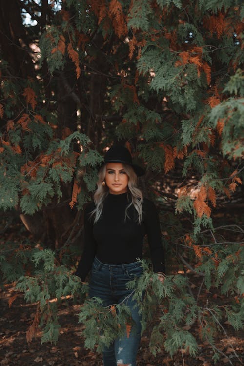 Trendy blond female in hat and casual outfit standing in trees with green and orange leaves