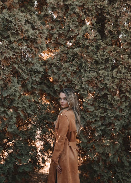 Side view of young female in elegant trench standing on background of green foliage in autumn park and looking at camera