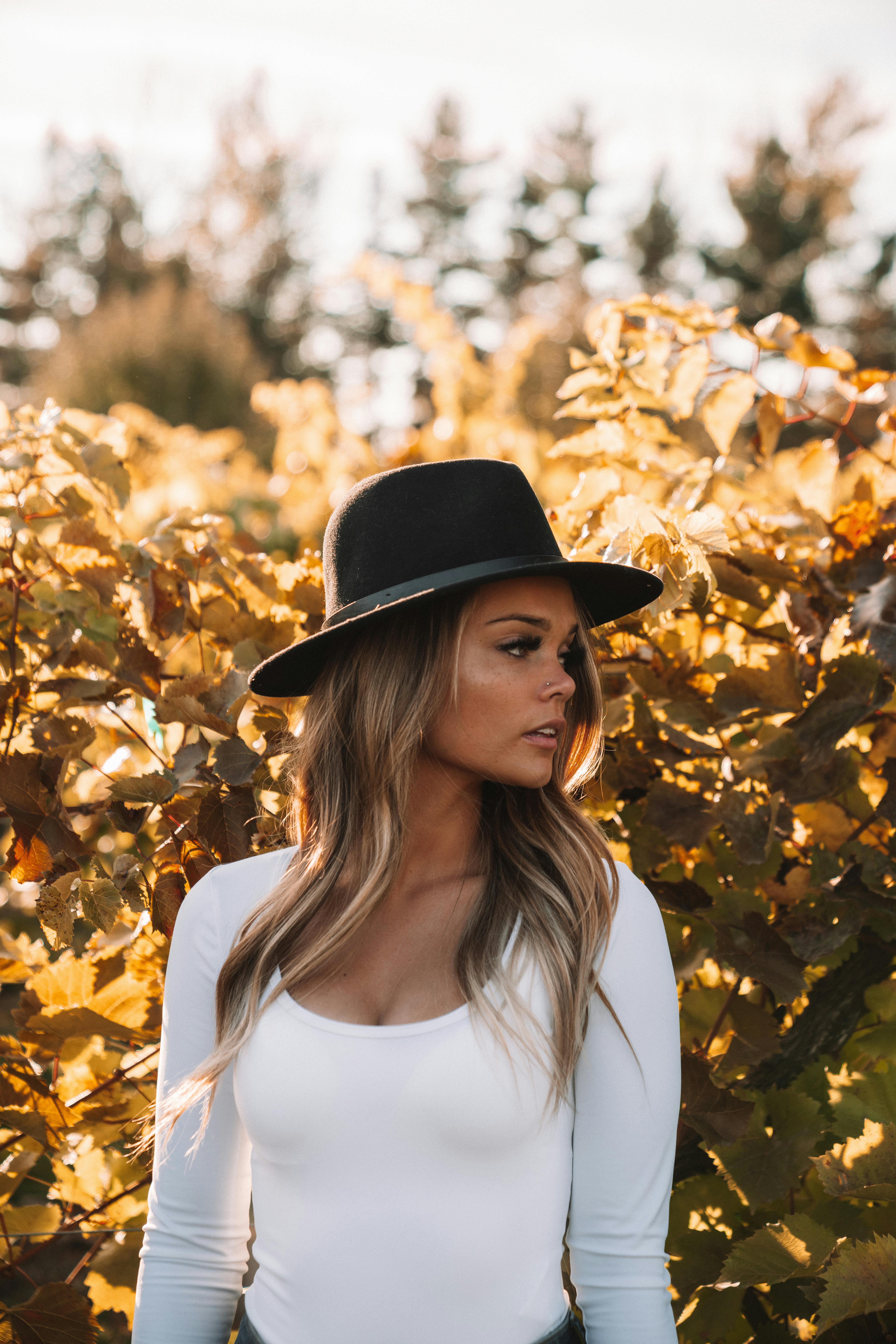 Trendy female with long hair wearing black hat and looking away against colorful yellow foliage