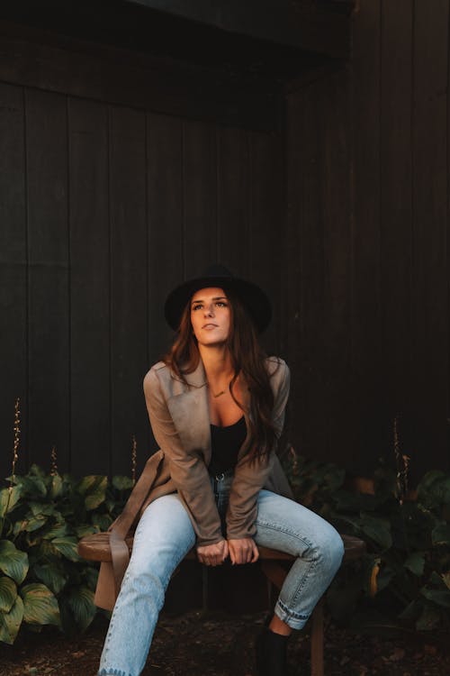 Full body of stylish young female in trendy outfit and hat sitting on wooden bench and looking away
