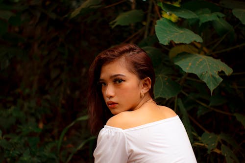 Side view of thoughtful young female millennial with long brown hair resting in lush green garden and looking at camera over shoulder