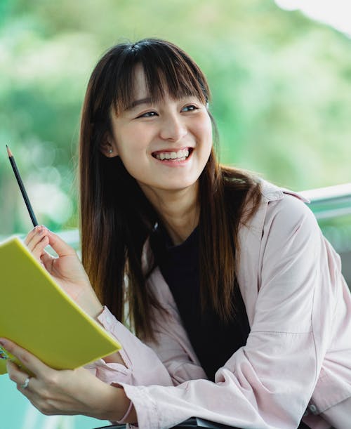 Happy ethnic female student in casual apparel with workbook and pencil looking away with toothy smile