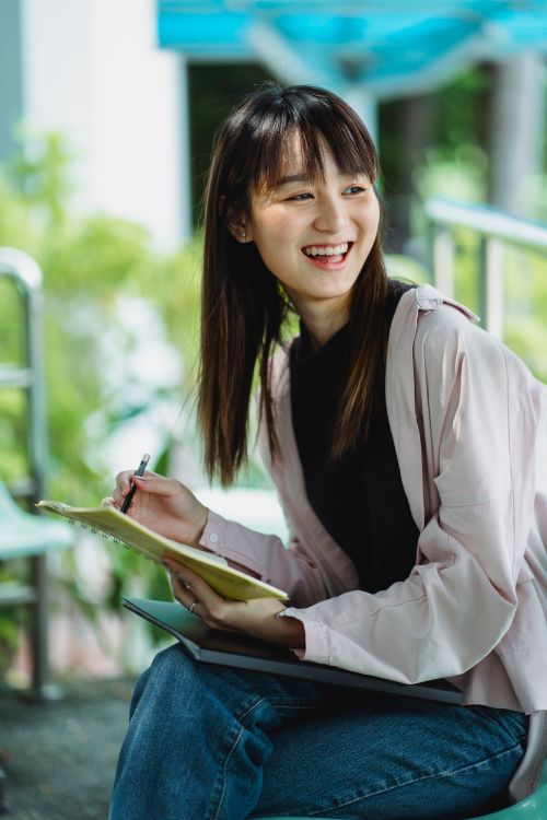 Free Cheerful ethnic female student with exercise book and pencil looking away while sitting with crossed legs Stock Photo