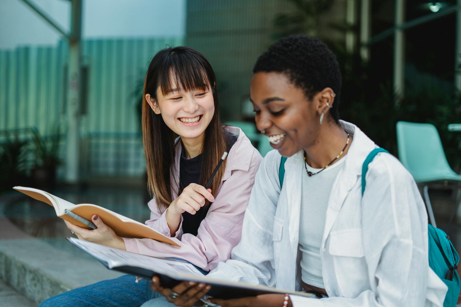 Young positive multiethnic female students with exercise books talking while doing homework on street stairs