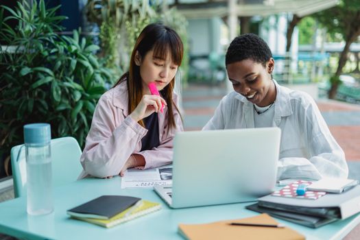 Two young women studying together at a table outdoors with a laptop and notebooks.