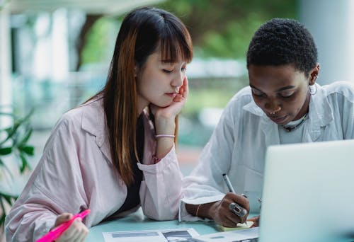 Young attentive Asian woman leaning on hand while studying with black girlfriend near netbook on street