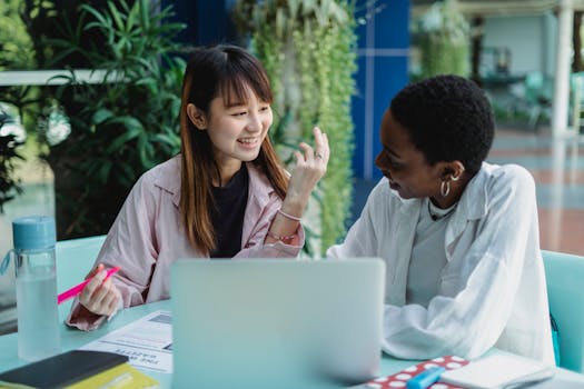 Young glad multiracial girlfriends talking while doing homework at table with netbook and looking at each other in garden