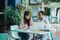 Young content diverse girlfriends talking while studying together at table with netbook in daylight