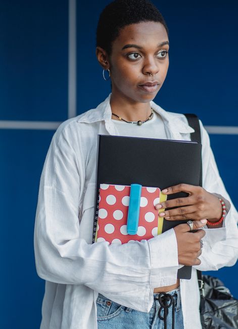 Crop young contemplative ethnic female in casual apparel with collection of exercise books looking away near wall