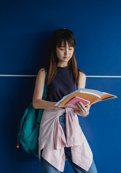 Free Young focused Asian lady with rucksack and assorted workbooks studying near blue wall in daytime Stock Photo