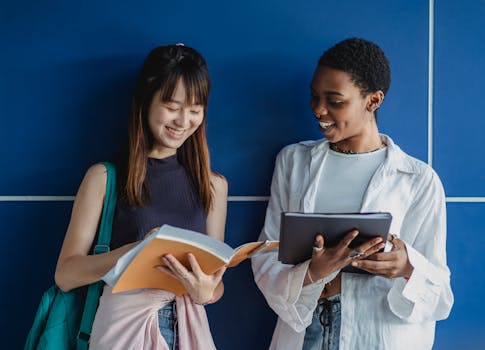 Two diverse young women smiling while studying together with books and tablets indoors.