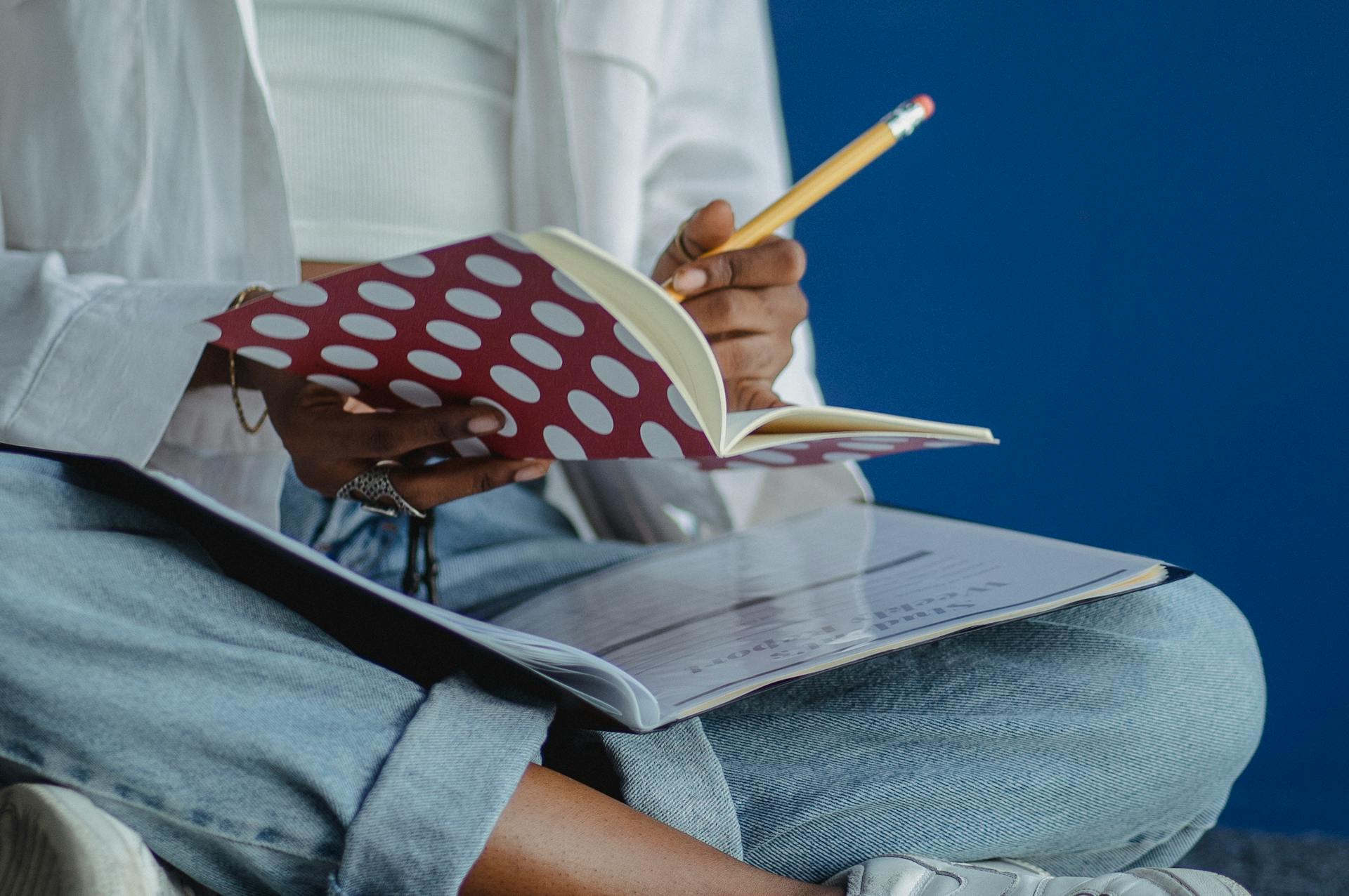 Crop anonymous diligent ethnic woman taking note with pencil in exercise book while studying with crossed legs on blue background