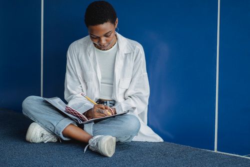 Free Young focused African American lady taking note in exercise book while studying with crossed legs on carpet covering Stock Photo