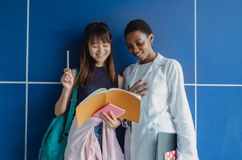 Young happy multiracial female students sharing workbook while studying together near blue wall in daytime