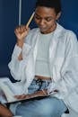 Crop young thoughtful African American female touching temple with pencil while watching exercise book and sitting with crossed legs near wall