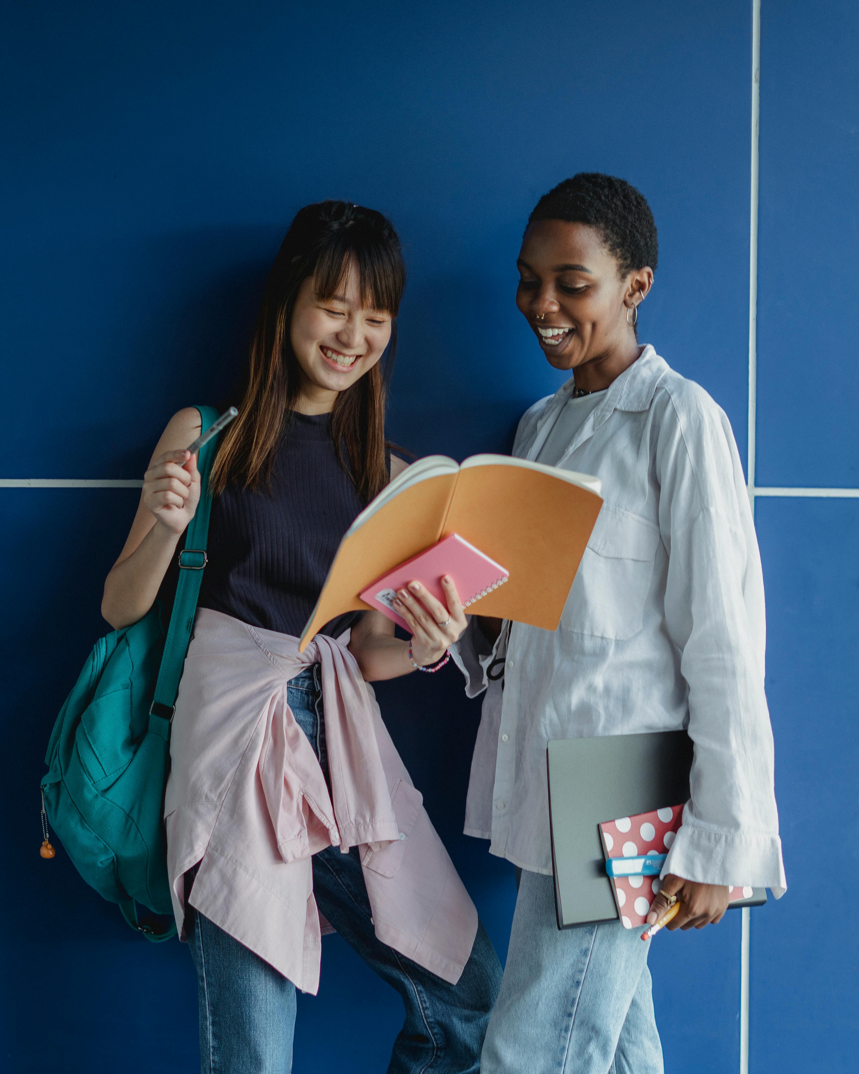 smiling multiracial girlfriends sharing workbook while doing homework