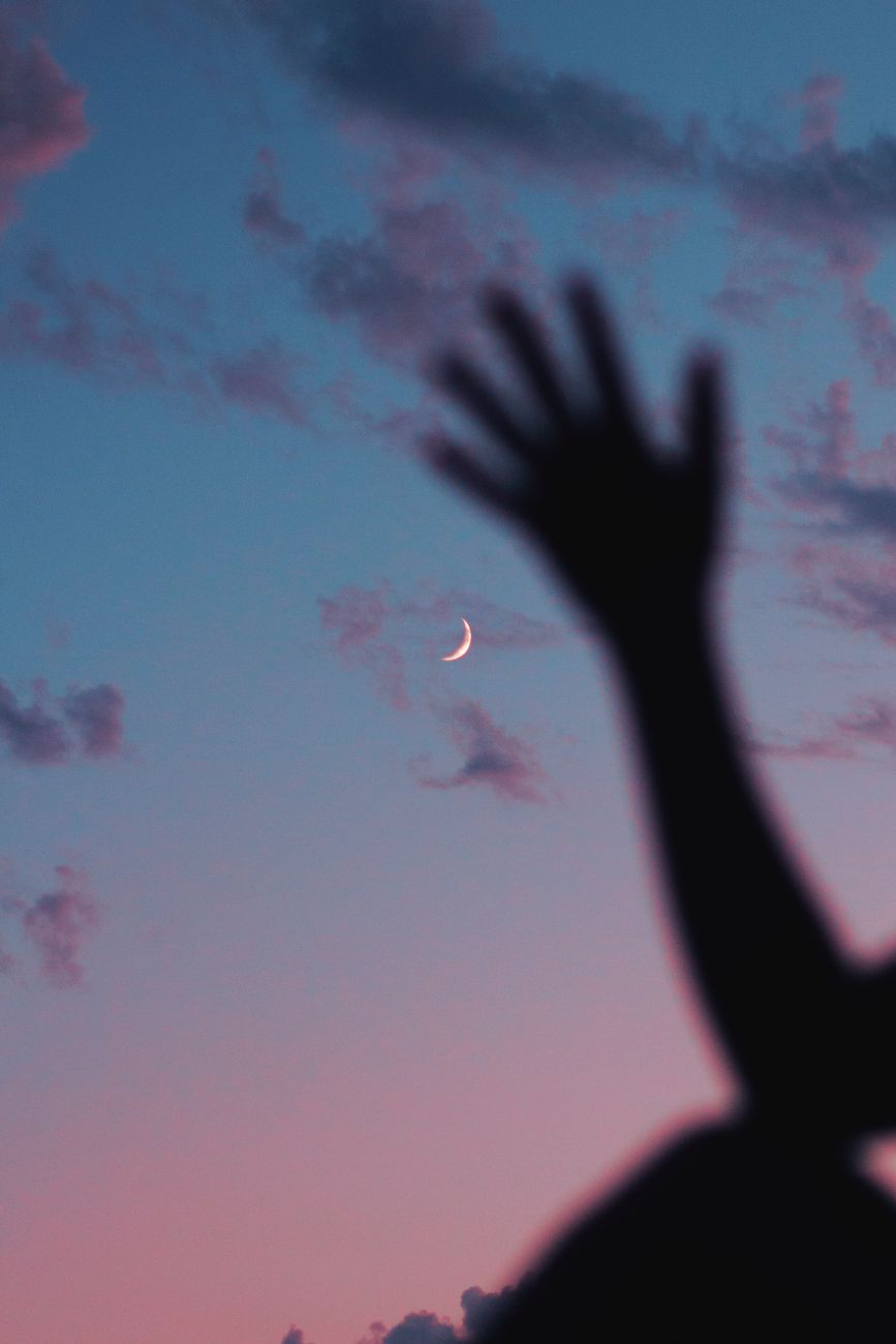 A silhouetted hand reaching towards a crescent moon in a twilight sky, creating a serene and dreamy scene.