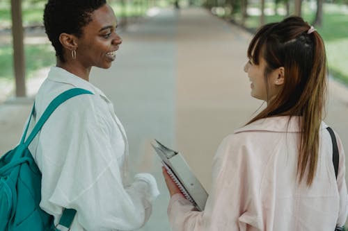 Back view of crop young smiling multiracial girlfriends with folder and rucksack looking at each other while conversing on walkway