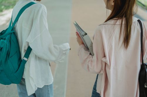 Free Back view crop female students in casual wear with copybooks walking together on sidewalk in campus and chatting Stock Photo