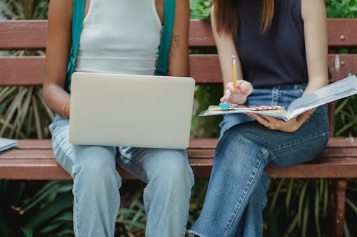 Crop anonymous diverse female students wearing casual outfit browsing netbook and taking notes in copybook while working on assignment on bench in park