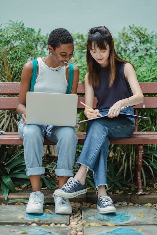 Full length content multiethnic female students in casual outfits browsing netbook and writing in workbook while preparing project together on bench in summer garden