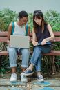 Full length content multiethnic female students in casual outfits browsing netbook and writing in workbook while preparing project together on bench in summer garden