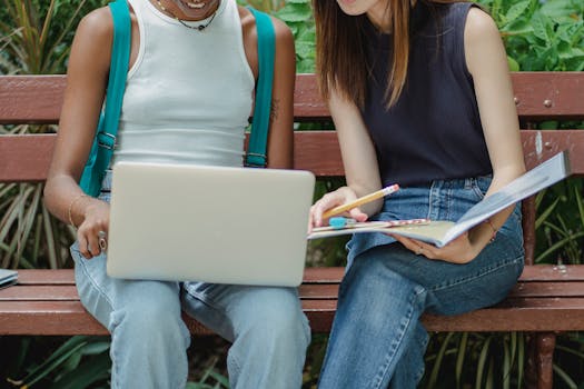 Crop multiethnic smiling female students in casual clothes browsing laptop and reading notes in copybook while sitting on bench in lush park