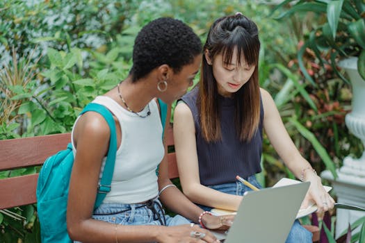 Two diverse female students studying together on a park bench with a laptop.