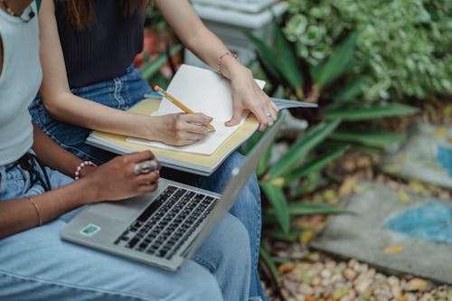 Free Crop faceless multiracial students in blue jeans using laptop and taking notes in workbook while preparing for exam together in lush summer park Stock Photo