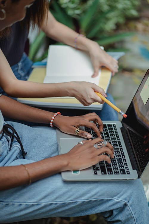 High angle side view anonymous multiracial girlfriends sitting together and using computer and notebook with pencil in daytime outside