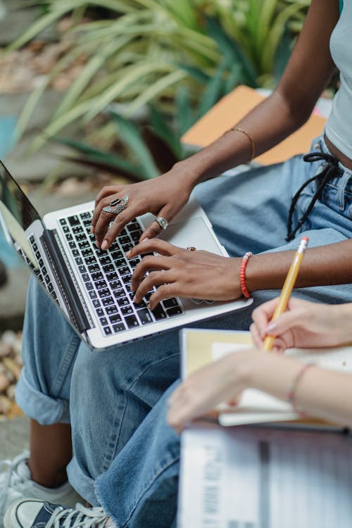 Free High angle crop faceless young multiracial women sitting in park with open laptop while writing with pencil in notepad Stock Photo