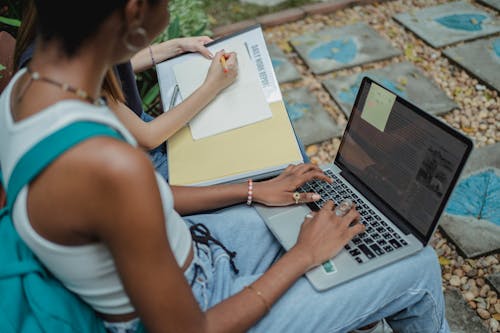 High angle anonymous young multiracial women sitting in yard while using computer and writing in notepad with pencil