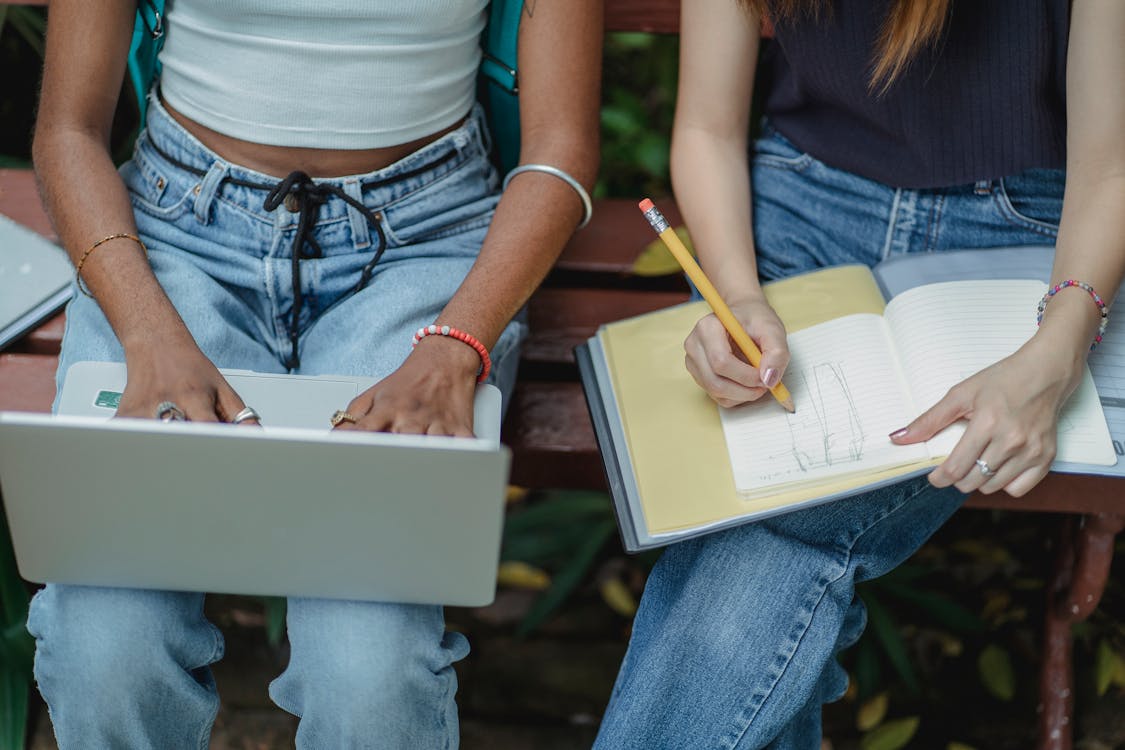 Free High angle crop faceless multiracial female friends typing on laptop and drawing in notebook with pencil while sitting on bench Stock Photo