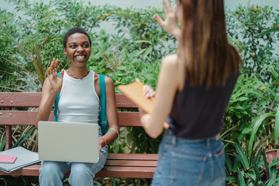 Smiling young multiracial women wearing casual clothes looking at each other while saying goodbye in daytime with netbook and notebooks