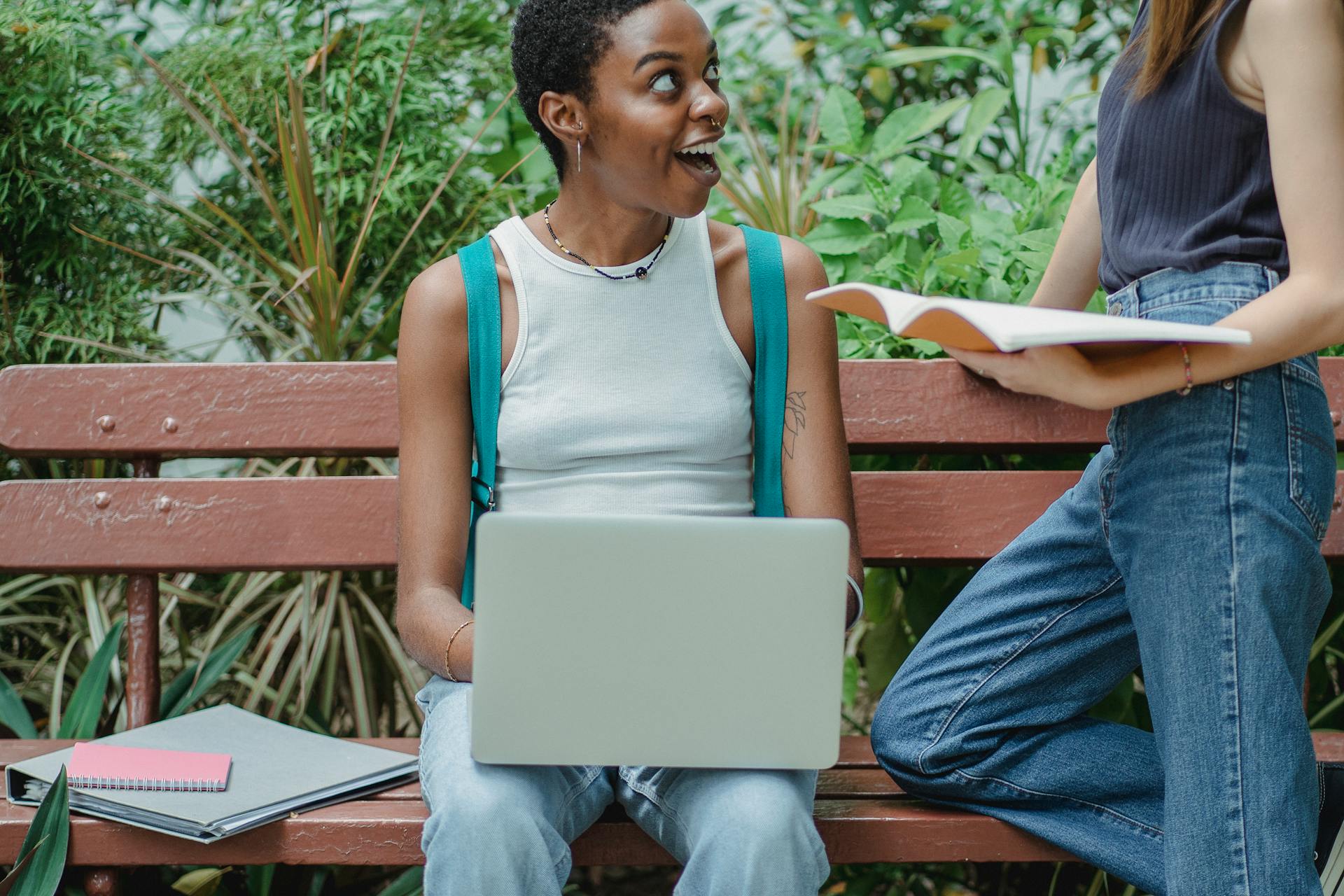 Multiracial female friends talking on bench in park while studying on laptop and using notepad sharing with exciting news