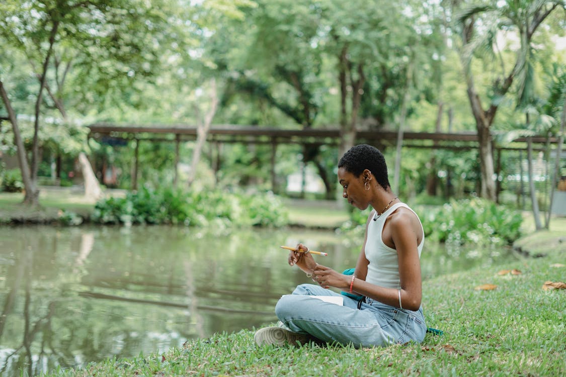 Free Side view of young African American female wearing casual clothes sitting near pond on green grass and using notepad with pencil in summer day Stock Photo