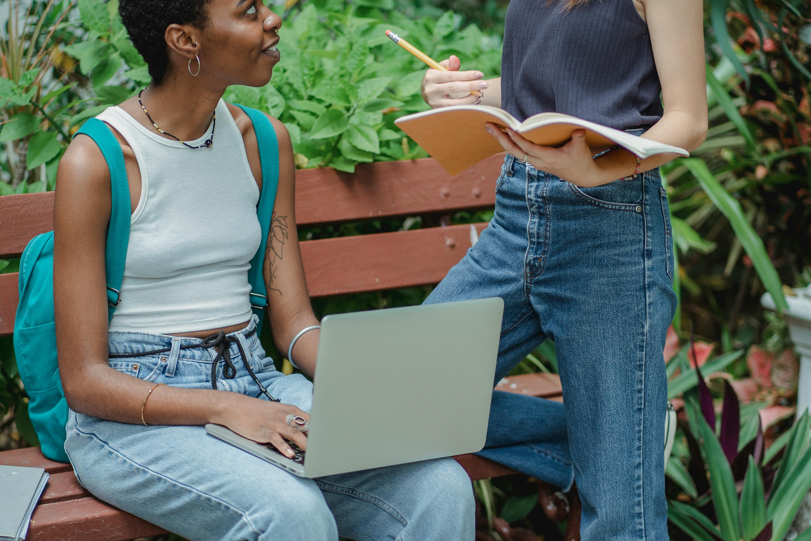 Multiracial women talking while using laptop and notebook on bench ...