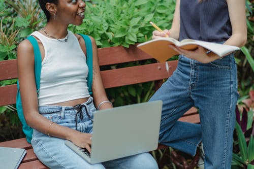 Crop multiracial female friends talking on bench in park while studying on computer and writing information in notebook