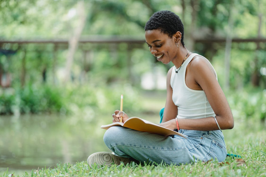 Black female writing in notepad while sitting on meadow