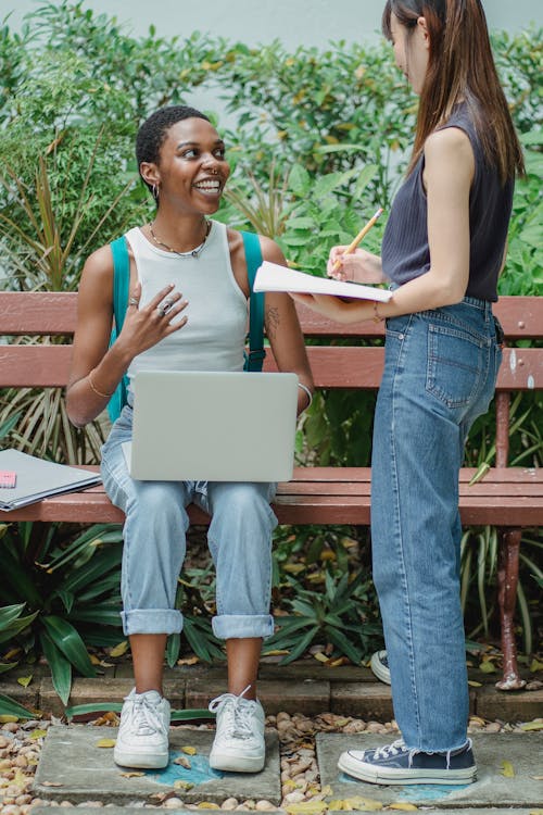 Full body multiracial female friends talking with each other outside while studying on netbook and using notebook while looking at each other